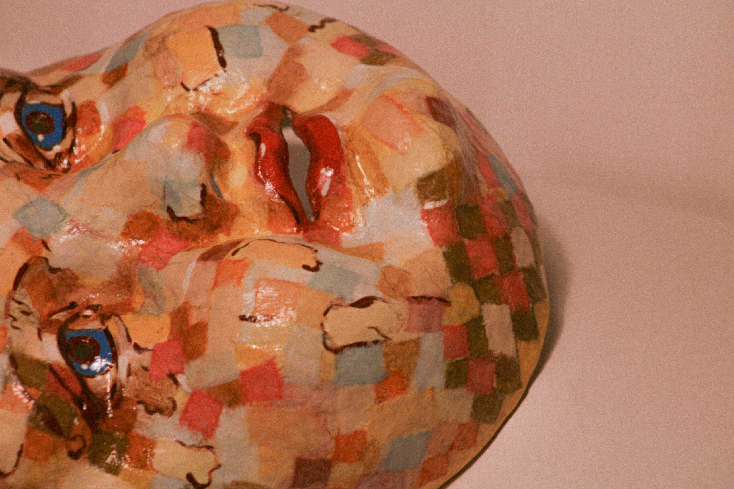 close-up of a sculpted face lying on its side, showing one blue eye, the nose bridge, and a mosaic of warm-toned painted squares with a glossy finish
