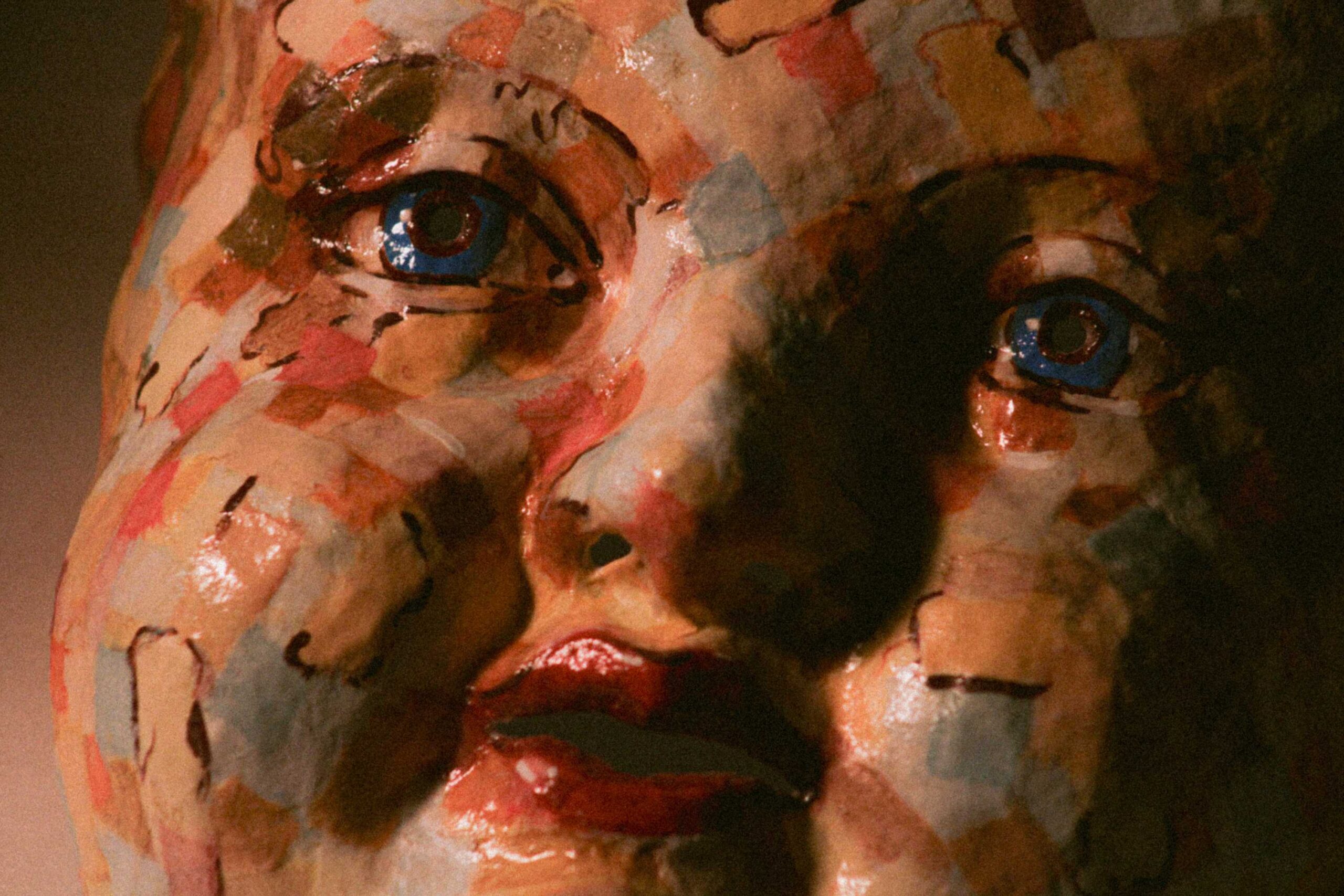 close-up of the eye area of the sculpture, showing blue eyes framed by painted brows and a patchwork surface under dramatic shadow
