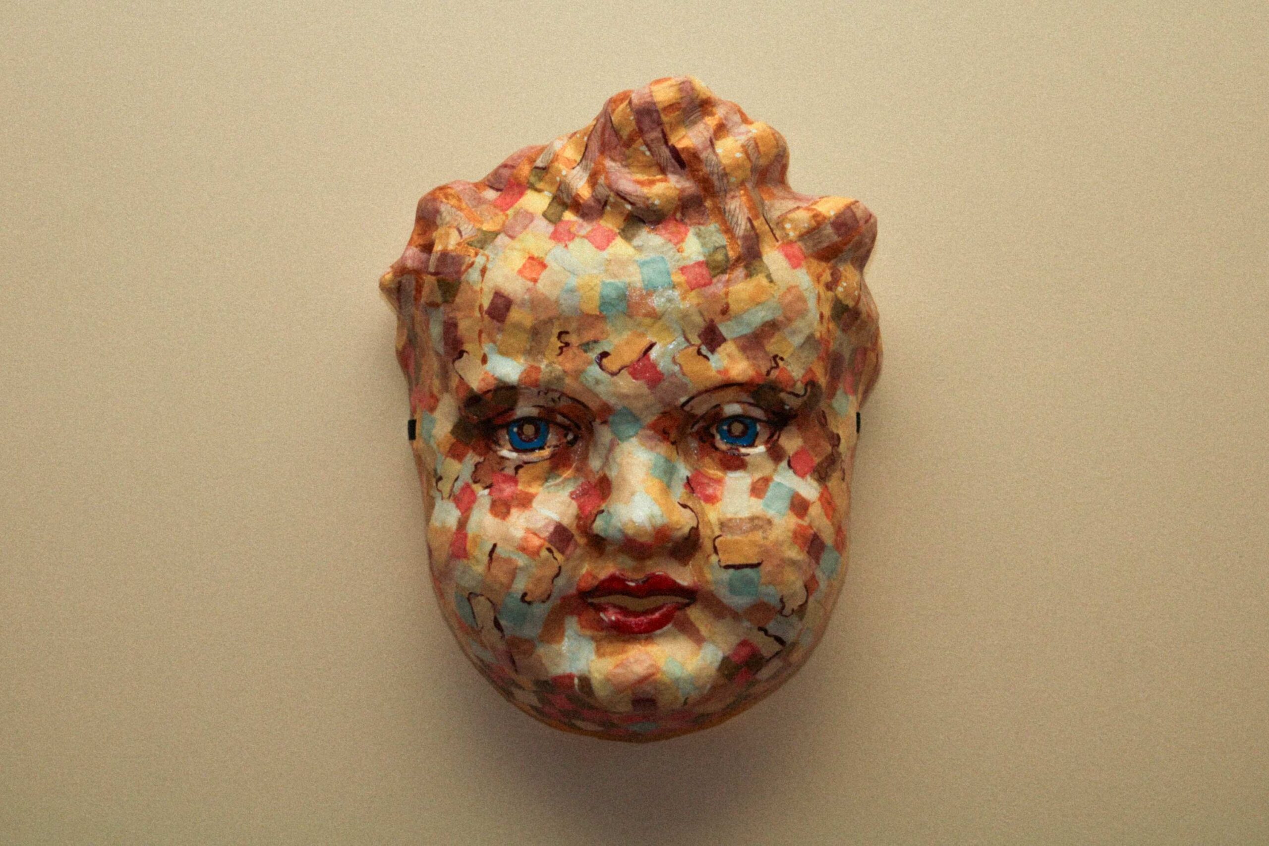 front view of the sculpted mask alone on a wall, evenly lit, showing the full face with blue eyes, red lips, and a dense mosaic of painted squares