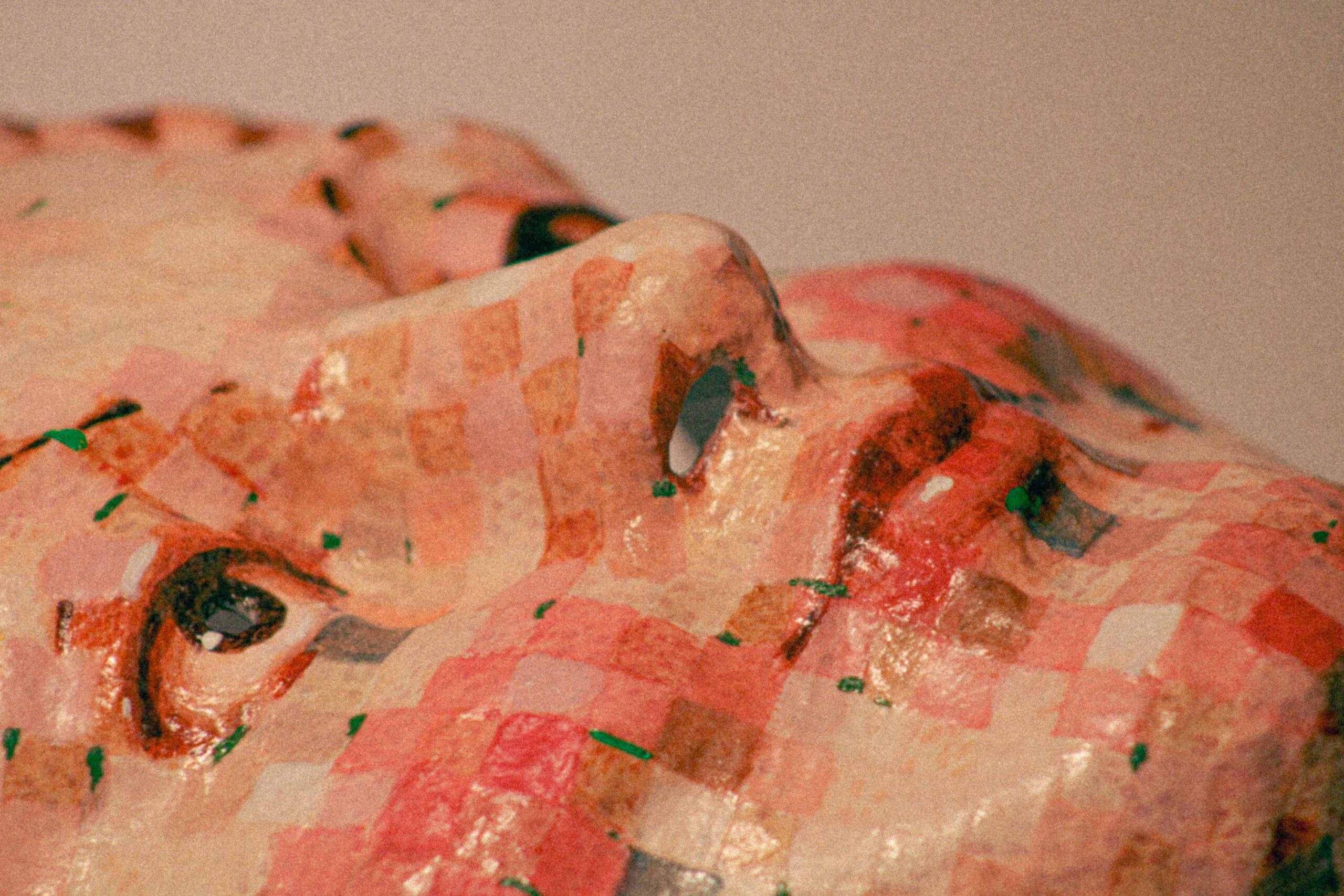 close-up of a sculpted face surface with a mosaic of pink, red and beige squares, showing part of an eye and nose with a glossy, handmade texture