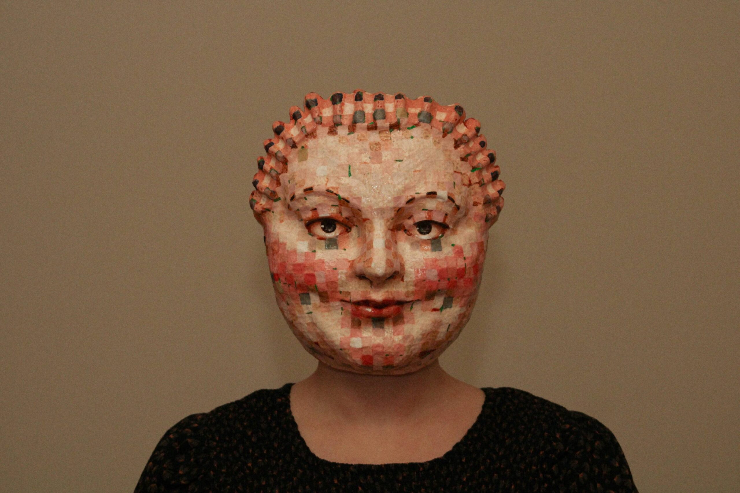 medium shot of a person wearing a mosaic-patterned sculptural mask, centered against a plain wall