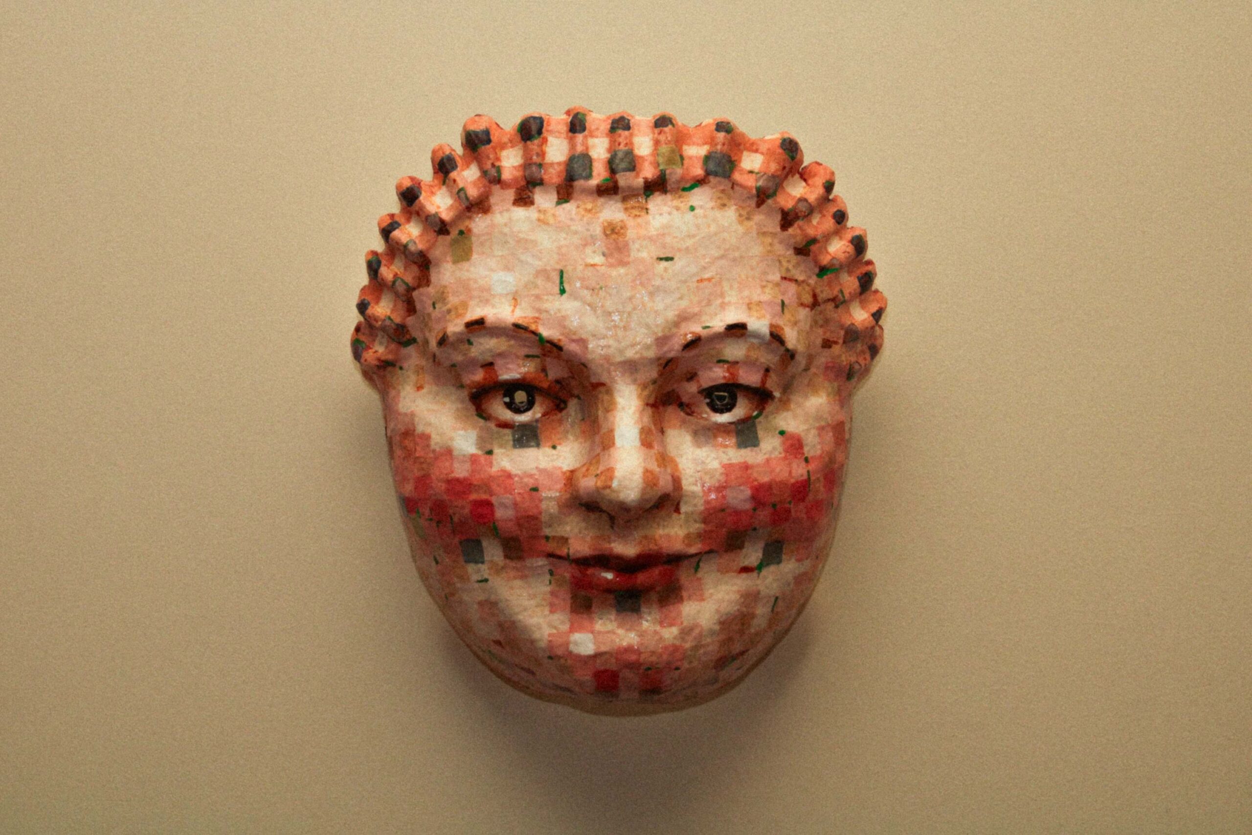front view of a mask-like sculpted face with symmetrical features, mosaic-style coloration, and a neutral expression against a plain background