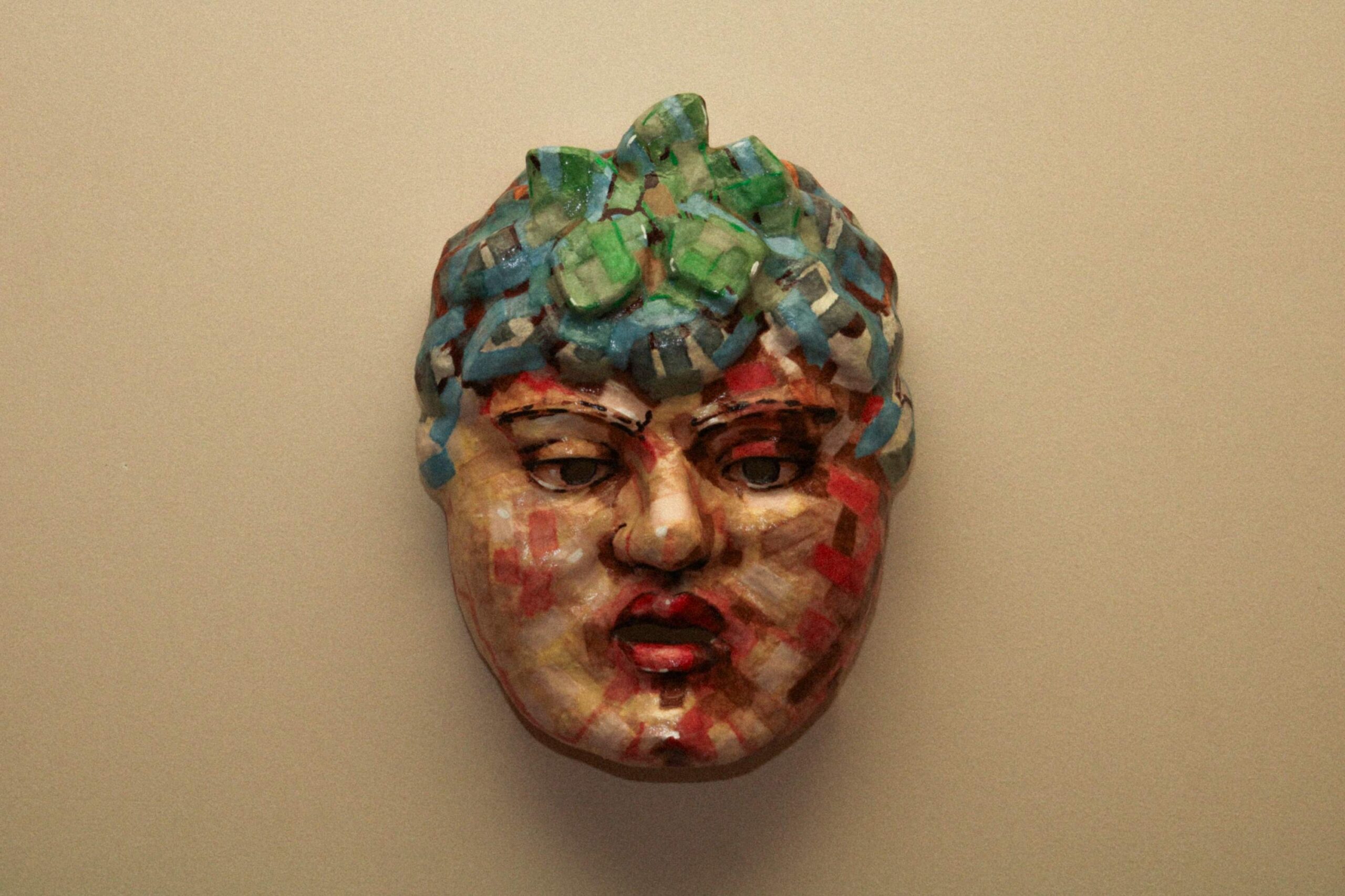 front view of the sculpted mask mounted on a wall, showing symmetrical facial features and multicolored mosaic-style painting