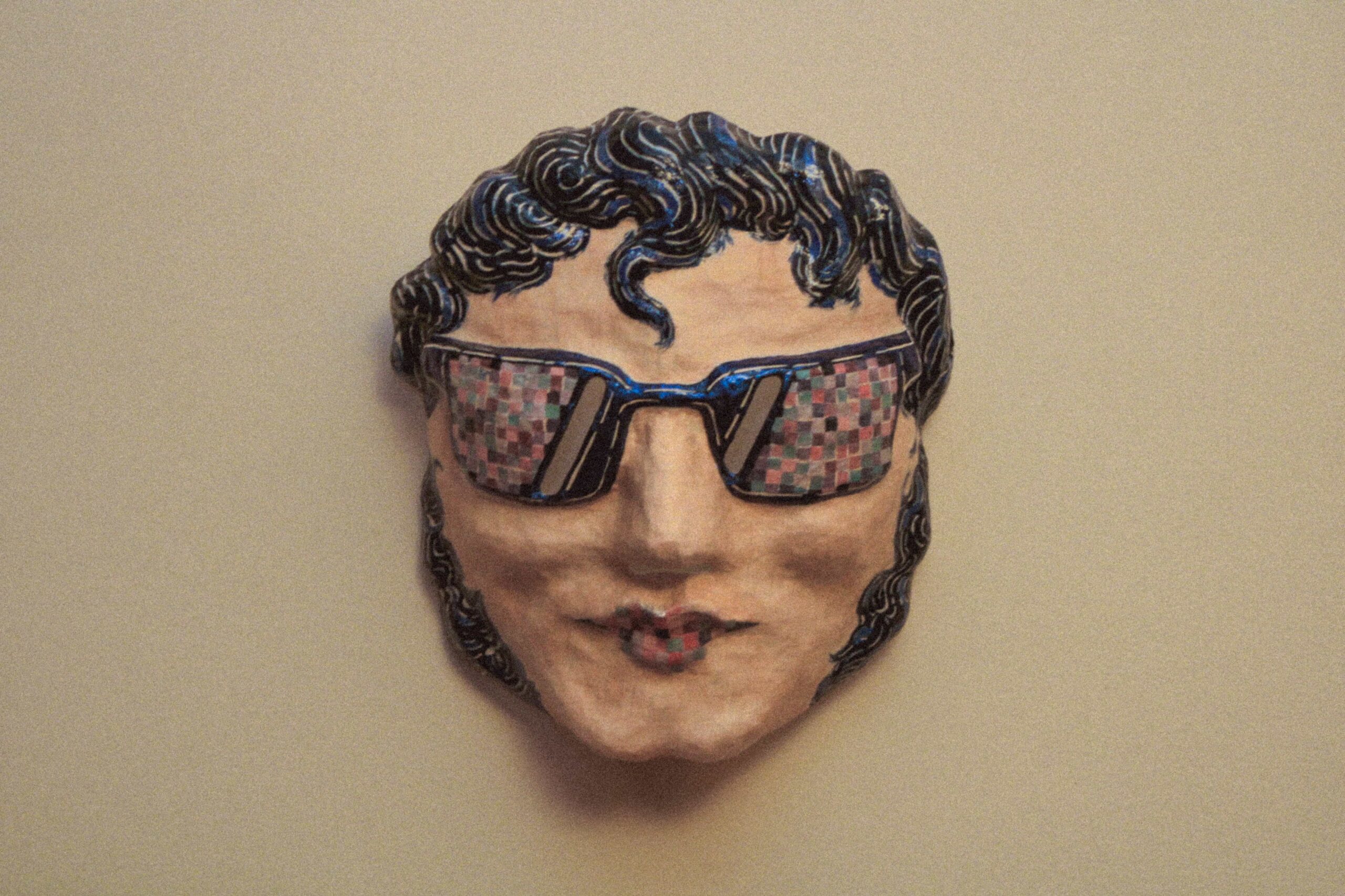 front view of a handmade contemporary art mask with sunglasses and stylized hair, mounted against a plain background