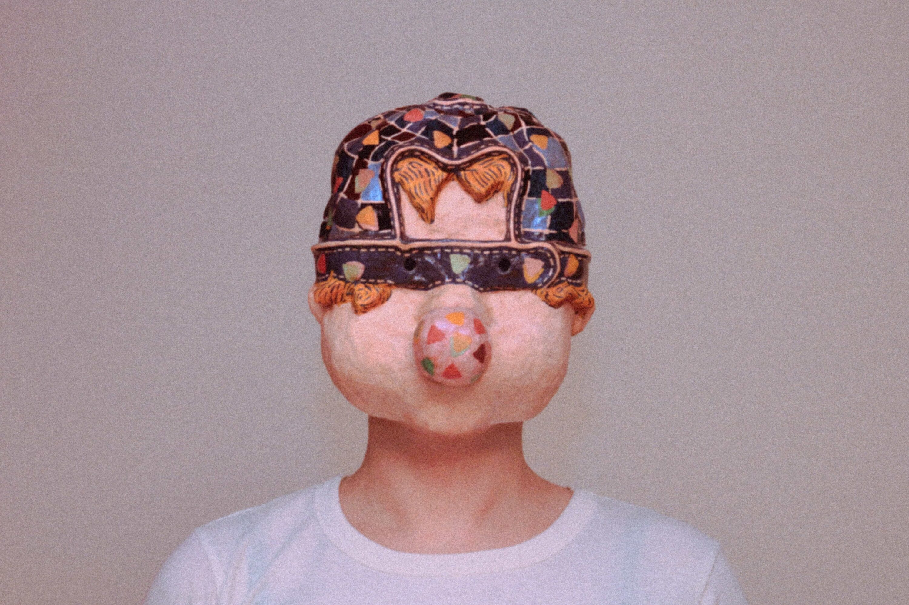 person wearing a handmade sculptural mask with mosaic pattern and protruding nose, against plain background